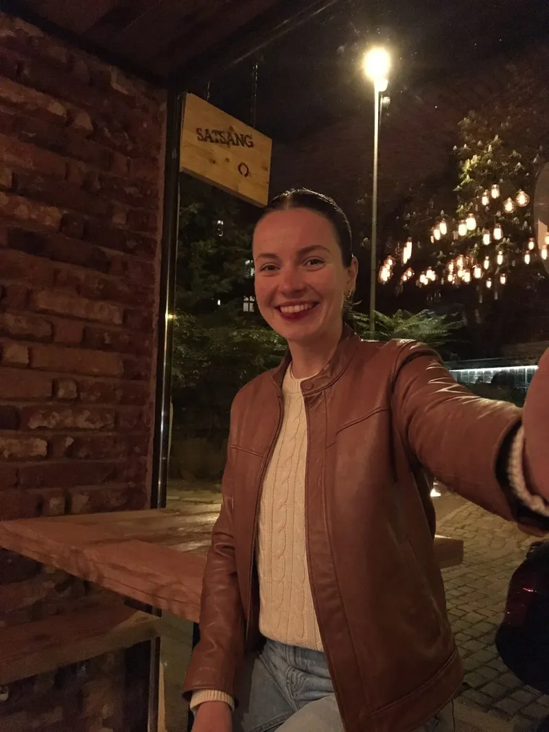 Accompanying digital illustration of Satsang’s historical interior: an added smiling figure in a brown jacket and light sweater, with a brick wall, wooden table and window bearing a SATSANG sign behind her. Not a photograph of a visit.
