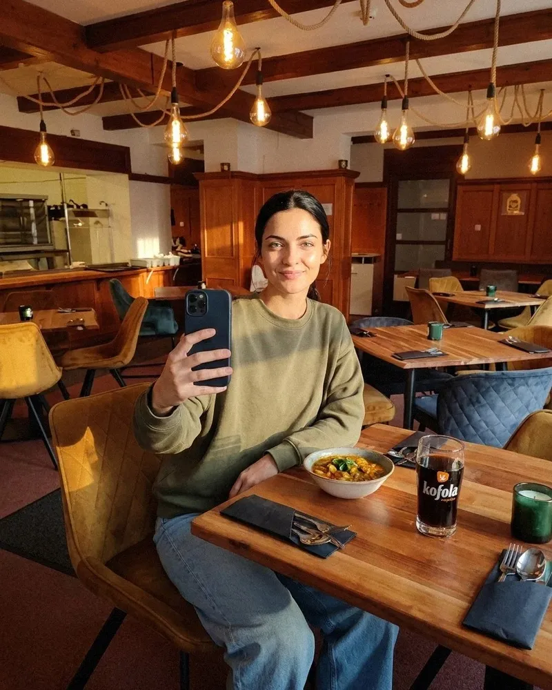 Tereza, wearing an olive sweatshirt and light blue jeans, sits at a wooden table holding a phone with its back towards the camera. In front of her are a bowl of food, a glass of Kofola and cutlery on a dark napkin. Colourful upholstered chairs, wood panelling and bulbs hanging from ropes are visible behind her.